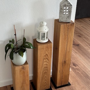 May include: Three wooden pedestal stands of varying heights, with a white lantern, a small concrete house, and a potted plant on top. The stands have black metal bases.
