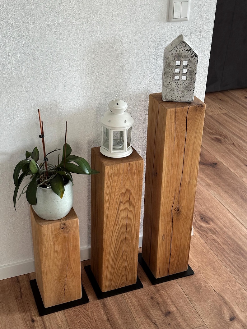 May include: Three wooden pedestal stands of varying heights, with a white lantern, a small concrete house, and a potted plant on top. The stands have black metal bases.