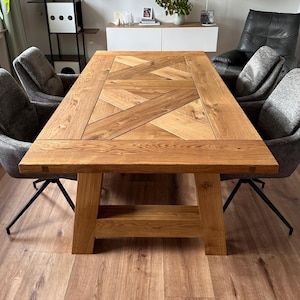 Könnte beinhalten: Ein rechteckiger Esstisch aus Holz mit hellbraunem Finish. Die Tischplatte hat ein Fischgrätenmuster. Der Tisch wird von dicken, stabilen Beinen getragen. Der Tisch ist von grau gepolsterten Stühlen mit schwarzen Metallbeinen umgeben. Der Tisch ist ca. 183 cm lang.