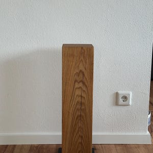 May include: A wooden pedestal with a natural wood finish and a black metal base. The pedestal is standing on a wooden floor.