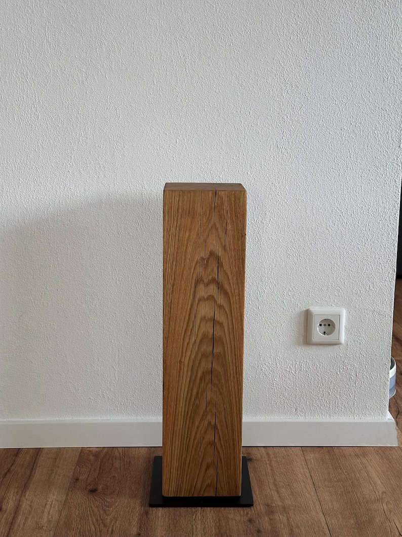 May include: A wooden pedestal with a natural wood finish and a black metal base. The pedestal is standing on a wooden floor.