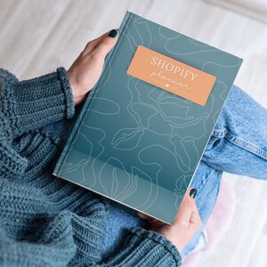 May include: A teal-colored Shopify planner with a floral design and the words "Shopify planner" on a peach-colored label. The planner is held in a person's lap, with the person wearing a blue sweater and jeans.