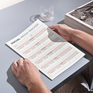 May include: A white Social Media Tracker sheet is held by a person. The sheet has pink boxes and the words "SOCIAL MEDIA TRACKER" at the top. A glass of water and a tray of glasses are in the background.