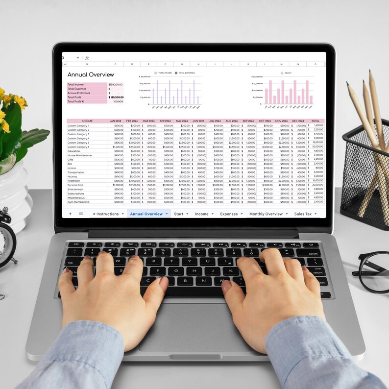May include: A laptop computer with a spreadsheet open on the screen. The spreadsheet shows an annual overview of income and expenses. The laptop is on a white desk with a person's hands typing on the keyboard.