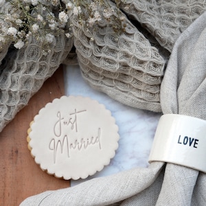 Könnte beinhalten: Ein weißer Serviettenring aus Keramik mit dem Wort "LOVE" darauf, neben einem weißen Keks mit den Worten "Just Married" in Prägung. Der Keks und der Serviettenring befinden sich auf einer weißen Oberfläche mit einem braunen und beigefarbenen, strukturierten Stoff im Hintergrund.