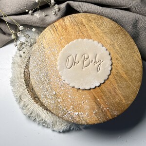May include: A round, light brown cookie dough stamp with the words "Oh Baby" embossed on it. The stamp is on a wooden surface with a white fringe around the edge.