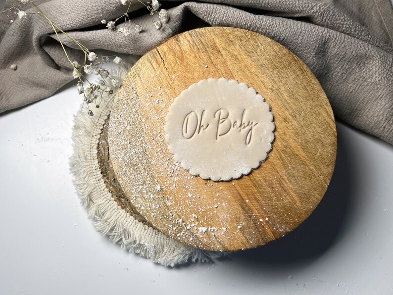 May include: A round, light brown cookie dough stamp with the words "Oh Baby" embossed on it. The stamp is on a wooden surface with a white fringe around the edge.