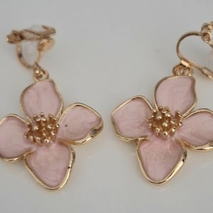 May include: A pair of clip-on earrings featuring a flower design. Each earring has a light pink enamel flower with gold-colored trim and a textured center. The clip-on mechanism is also gold-colored.