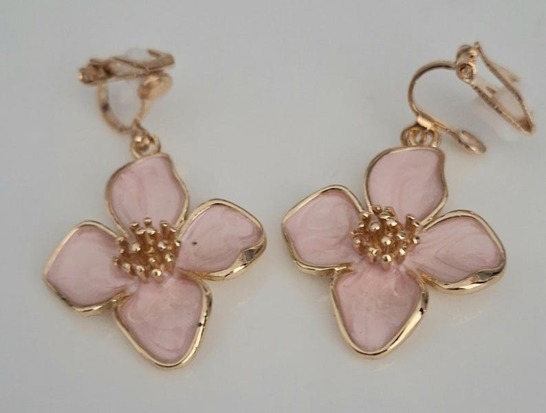 May include: A pair of clip-on earrings featuring a flower design. Each earring has a light pink enamel flower with gold-colored trim and a textured center. The clip-on mechanism is also gold-colored.