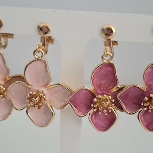 May include: Two pairs of clip-on earrings featuring flower designs. One pair is light pink, the other is a deeper pink. Each flower has a gold-colored center and a gold-colored border. The earrings are displayed on a white stand.