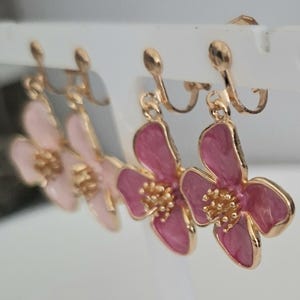May include: A set of four flower-shaped clip-on earrings. The earrings feature a gold-colored metal frame with pink and light pink enamel petals and a gold-colored center. The earrings are displayed on a white earring holder.