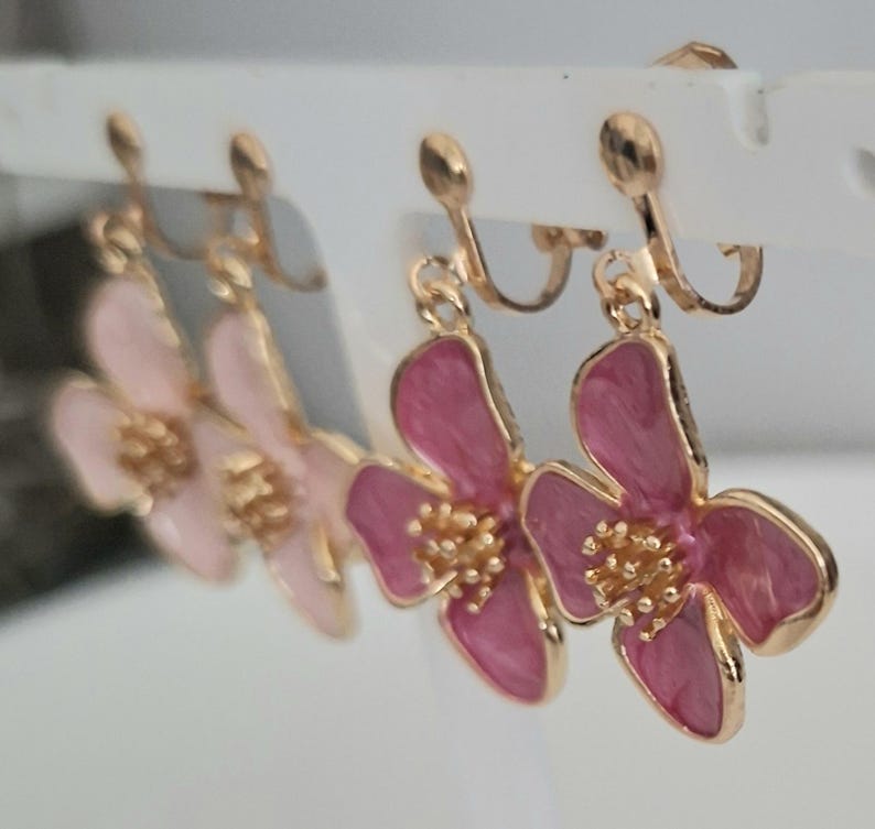 May include: A set of four flower-shaped clip-on earrings. The earrings feature a gold-colored metal frame with pink and light pink enamel petals and a gold-colored center. The earrings are displayed on a white earring holder.