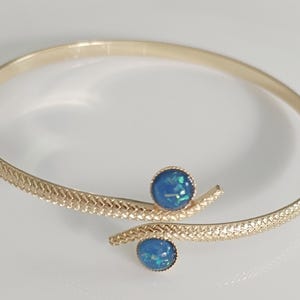 Könnte beinhalten: Ein goldenes Armreif-Armband mit einem strukturierten Schlangendesign und zwei blauen Opal-Steinen.