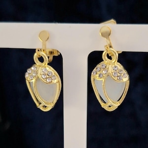 May include: Gold-coloured clip-on earrings. Each earring has a teardrop shape with a white centre and a top section decorated with small, clear crystals. The earrings are displayed on a white stand against a dark blue background.