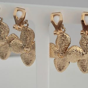 May include: Gold-toned clip-on earrings. Each earring features a textured, flower-shaped design with four petals. The clip-on mechanism has a white, rectangular accent. The earrings are displayed on a white stand.