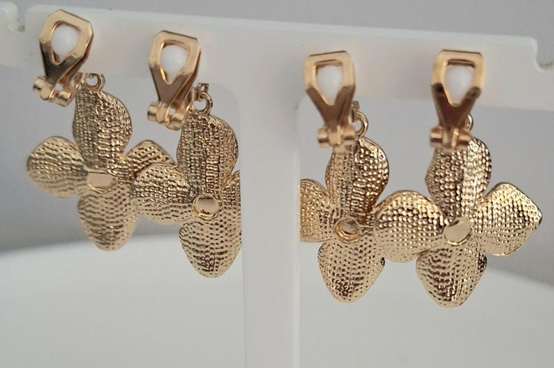 May include: Gold-toned clip-on earrings. Each earring features a textured, flower-shaped design with four petals. The clip-on mechanism has a white, rectangular accent. The earrings are displayed on a white stand.