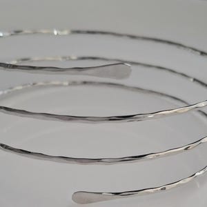 Puede incluir: Un brazalete plateado con textura martillada. El brazalete se enrolla alrededor del brazo, con un extremo plano y redondeado. La joya se exhibe sobre una superficie blanca. El brazalete es una pieza de joyería de moda.