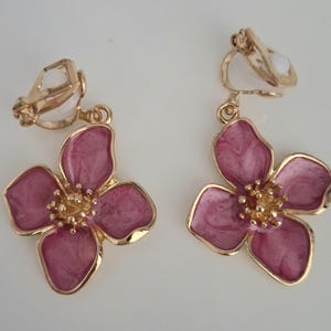 May include: Clip-on earrings featuring a flower design. The earrings have a gold-colored metal frame and a pink enamel flower with a gold-colored center. The flower petals have a marbled effect. The earrings are designed to clip onto the ear.
