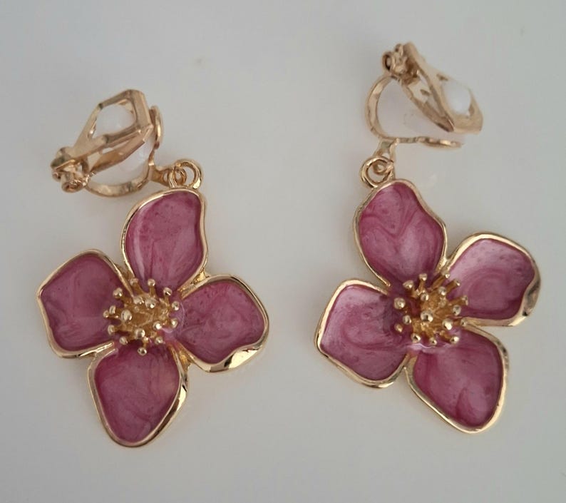 May include: Clip-on earrings featuring a flower design. The earrings have a gold-colored metal frame and a pink enamel flower with a gold-colored center. The flower petals have a marbled effect. The earrings are designed to clip onto the ear.