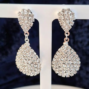 May include: A pair of silver-toned drop earrings featuring a teardrop-shaped pendant covered in small, clear, faceted crystals. Each earring has a smaller, triangular-shaped crystal-encrusted stud at the top, connected by a small silver ring.