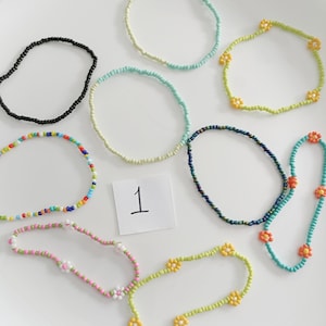 Puede incluir: Surtido de pulseras de cuentas coloridas con detalles de flores. Algunas pulseras son de colores sólidos, mientras que otras tienen cuentas multicolores. Las pulseras están dispuestas sobre una superficie blanca con un número "1" escrito a mano en una tarjeta blanca.