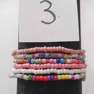 Può includere: Un set di otto braccialetti colorati con perline, con un design rosa, bianco e multicolore. I braccialetti sono esposti su una superficie nera con un'etichetta bianca che dice "3."