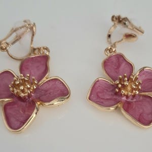 May include: A pair of clip-on earrings featuring a flower design. The earrings have a gold-colored metal frame and a pink enamel finish. The flower petals are a deep pink with a marbled effect, and the center is a textured gold color.
