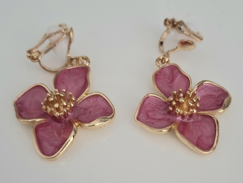 May include: A pair of clip-on earrings featuring a flower design. The earrings have a gold-colored metal frame and a pink enamel finish. The flower petals are a deep pink with a marbled effect, and the center is a textured gold color.