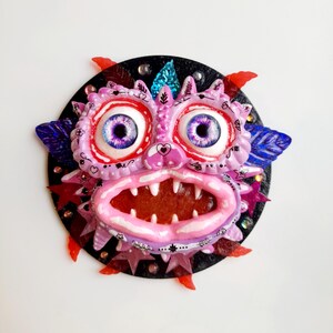 Puede incluir: Una cara de monstruo rosa y morado con ojos grandes, una boca abierta de par en par con dientes afilados y un fondo negro. El monstruo está decorado con purpurina y estrellas.