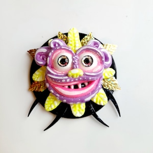 Puede incluir: Una cara de monstruo púrpura y rosa con detalles amarillos y blancos, ojos grandes y una amplia sonrisa. El monstruo está rodeado de hojas doradas y púas negras.