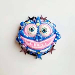 Puede incluir: Un monstruo sonriente azul y rosa con ojos saltones y un fondo brillante. El monstruo tiene una boca rosa con dientes blancos y está rodeado de estrellas negras.