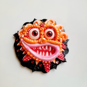 Puede incluir: Una escultura de arcilla de color naranja brillante y rosa de una cara de monstruo con ojos rojos y una sonrisa de dientes. La escultura está rodeada de un fondo negro con pequeños detalles negros y rojos.