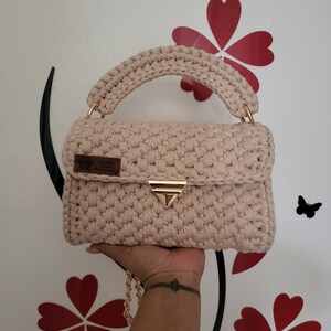 May include: A light pink crocheted handbag with a gold chain strap and a gold clasp. The bag has a brown leather tag with the word "remarkable" on it.
