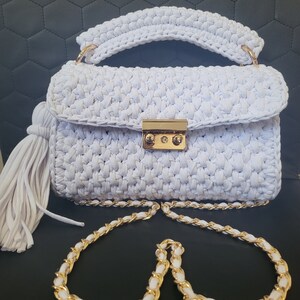 May include: A white crocheted handbag with a gold chain strap and a tassel. The bag has a gold clasp closure.