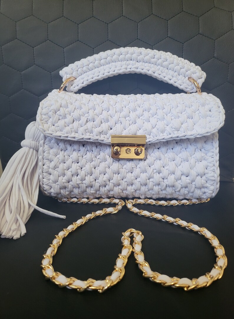 May include: A white crocheted handbag with a gold chain strap and a tassel. The bag has a gold clasp closure.