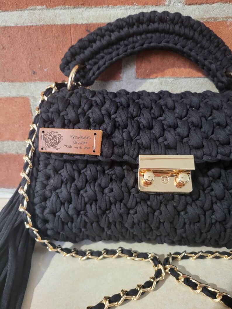 May include: Black crocheted handbag with a gold chain strap and a brown leather tag that reads "Frankly's Crochet Made with Love".
