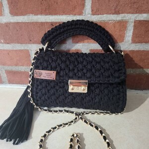 May include: Black crocheted handbag with a gold chain strap and a gold lock closure. The bag has a black tassel and a small brown leather tag that says "Frankie's Crochet Made with Love".