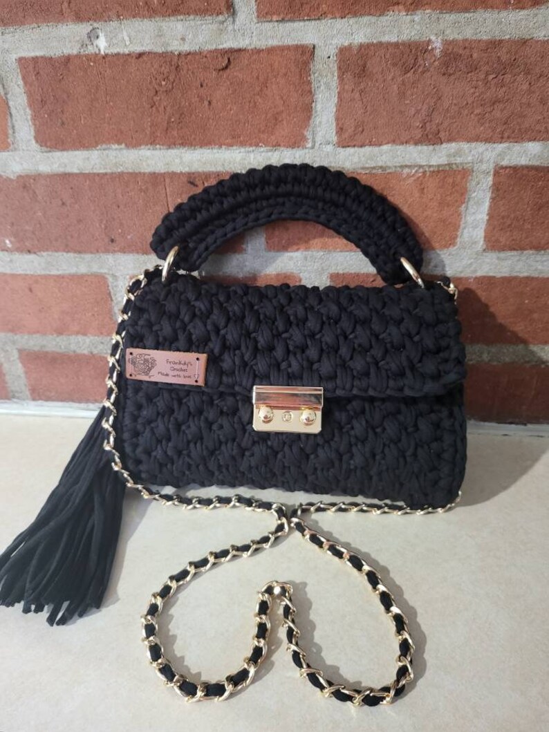 May include: Black crocheted handbag with a gold chain strap and a gold lock closure. The bag has a black tassel and a small brown leather tag that says "Frankie's Crochet Made with Love".