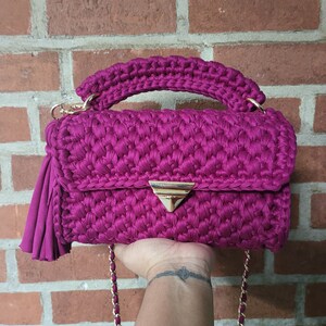 May include: A magenta crocheted handbag with a gold chain strap and a gold triangular clasp. The bag has a tassel on one side.