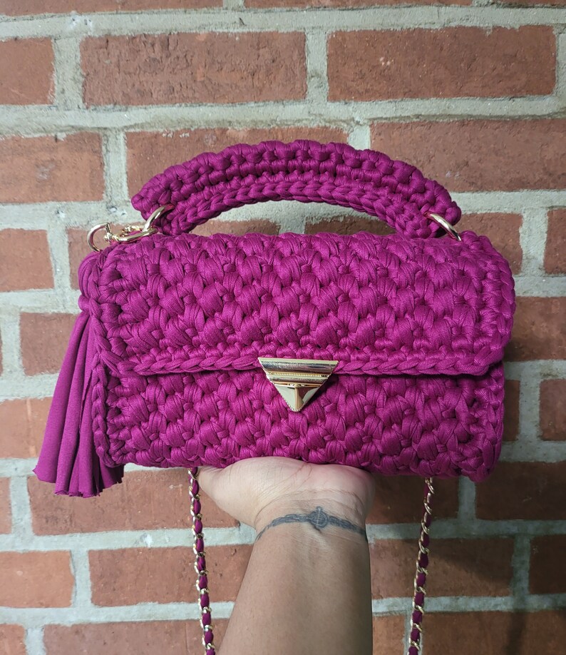 May include: A magenta crocheted handbag with a gold chain strap and a gold triangular clasp. The bag has a tassel on one side.
