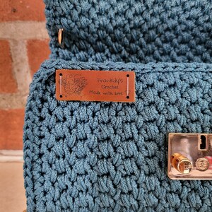 May include: A teal blue crocheted handbag with a brown leather tag that reads "Frankidy's Crochet Made with love". The bag has a gold metal clasp.
