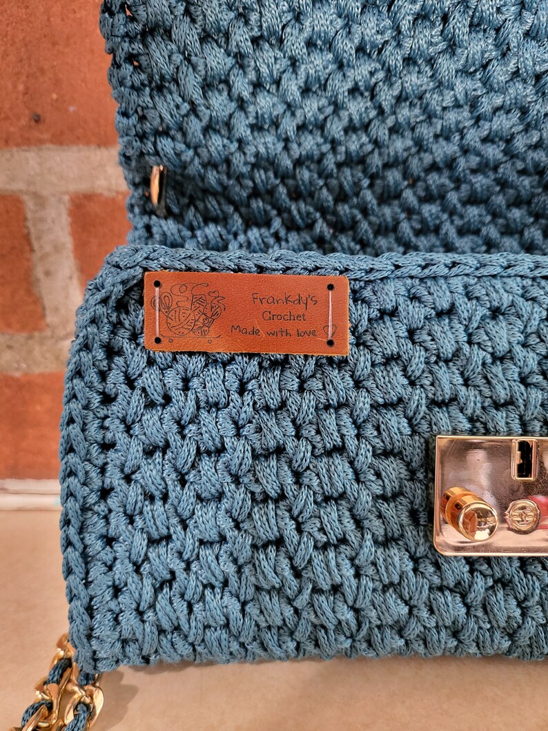 May include: A teal blue crocheted handbag with a brown leather tag that reads "Frankidy's Crochet Made with love". The bag has a gold metal clasp.