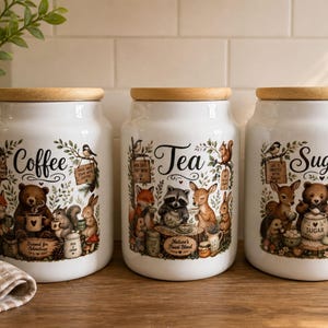 May include: Three white ceramic canisters with wooden lids, labelled "Coffee," "Tea," and "Sugar." Each canister features a woodland animal illustration with text. The canisters are on a wooden surface.