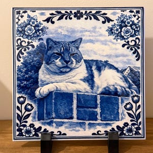 May include: Square ceramic tile with a blue and white design. The tile depicts a tabby cat resting on a brick wall, with a floral border. The background shows a sky and trees. The tile is displayed on a black stand.