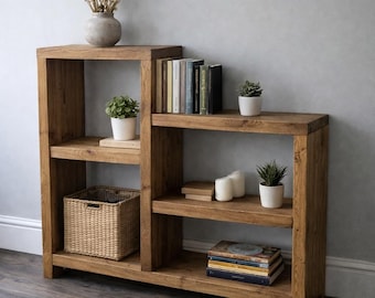 May include: A tiered wooden bookshelf with a natural brown finish, featuring a tall left section and a lower right section. The shelves hold a woven basket, stacks of books, three small potted green plants, and a vase with dried flowers.