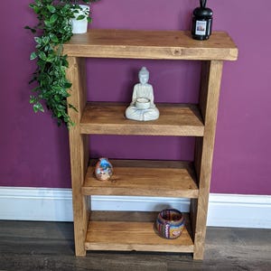 May include: A three-tier wooden bookshelf with a medium brown finish. The top shelf holds a green leafy plant in a white pot and a black reed diffuser. The middle shelf features a light grey Buddha figurine. The bottom shelf contains two small decorative bowls.