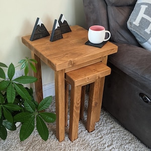 May include: A set of two wooden nesting tables with a natural wood finish. The tables are square and have a simple design. The top table has a cup and saucer on it, and the bottom table has a small wooden mountain sculpture on it.