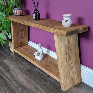 May include: A wooden console table with a lower shelf, stained a warm brown. The table has a simple, modern design. Decorative items, including a small Buddha statue, are displayed on the shelves. The table is 152 cm long.