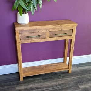 May include: A wooden console table with two drawers and a lower shelf. The table has a natural wood finish and brass drawer pulls. The table is in front of a purple wall and a potted plant.