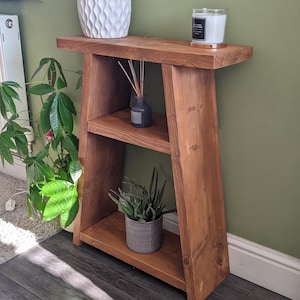 May include: A wooden A-frame shelf with two shelves. The shelf is stained a dark brown colour and has a white ceramic vase with a textured design on the top shelf. A candle and a diffuser are also on the top shelf. A potted succulent plant is on the bottom shelf.